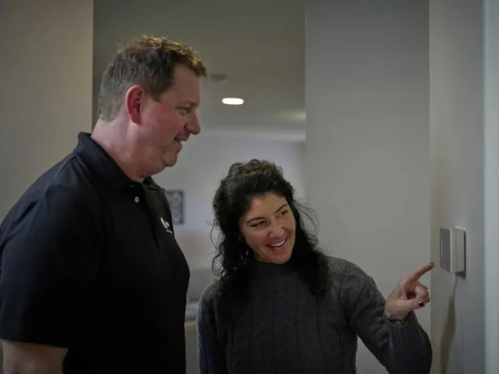 Woman pointing to digital thermostat on her wall with an HVAC technician standing beside her.