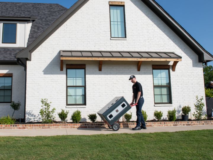 An HVAC technician using a two-wheeled dollie to transport a new Trane furnace along a walkway outside a white brick house.