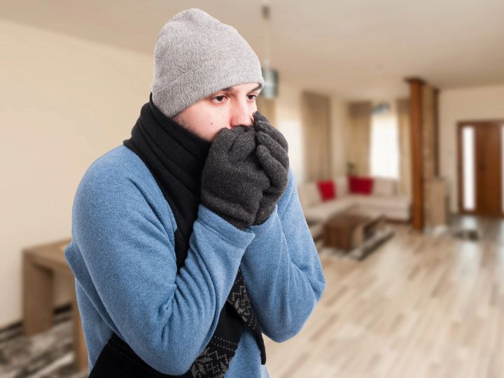 A man indoors wearing a hat, scarf and gloves, shivering and blowing into his hands to stay warm in a chilly room.