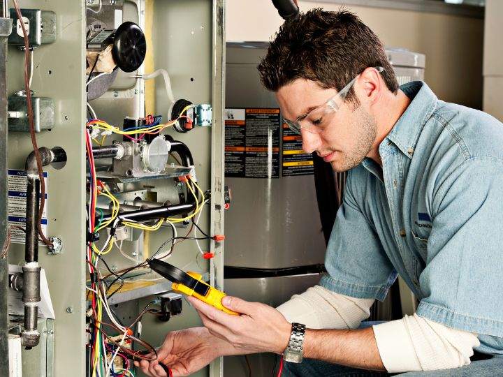 An HVAC technician wearing safety glasses uses a yellow digital multimeter to check the electrical wiring inside a furnace panel.