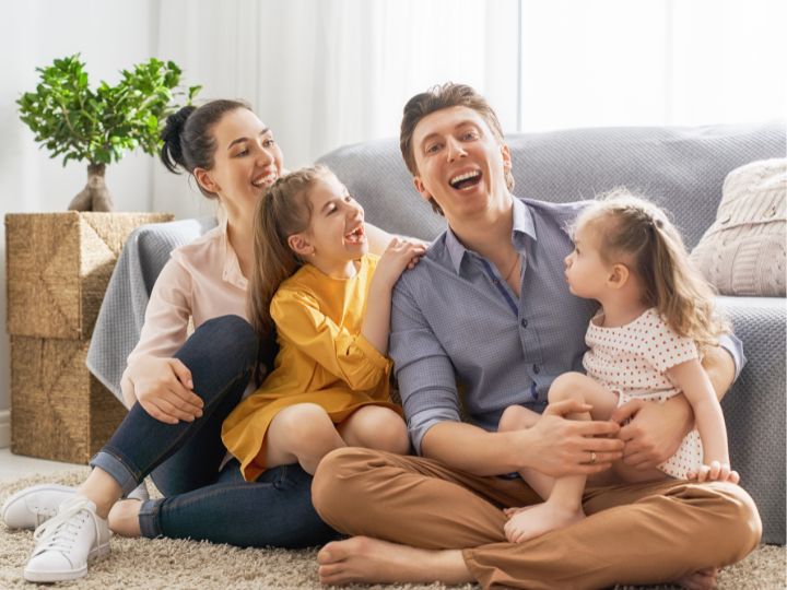 Smiling parents spending time with their two young daughters in their living room.